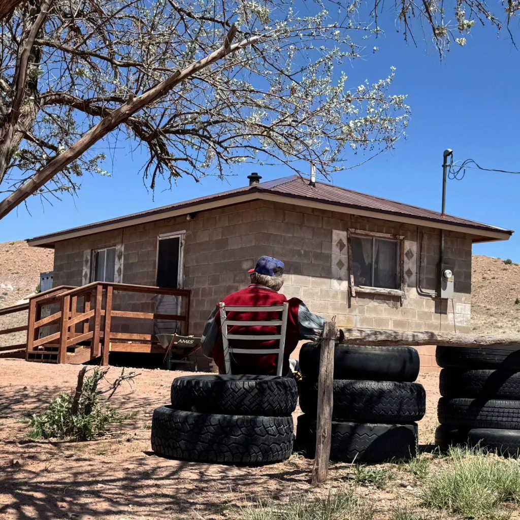 An elderly Navajo man, Charley Billy, sits outside his home, which recently received renovations through Yee Ha'ólníi Doo's 'Owner-Occupied Housing Health and Safety' initiative. The improvements include a newly built access ramp and a new roof. Previously, the homeowner struggled with unsafe steps and lacked a handrail, leading to a fall last year, and relied on neighbor assistance for roof repairs. The initiative is funded by the Arizona Department of Housing, providing critical support to vulnerable households in Northern Arizona.