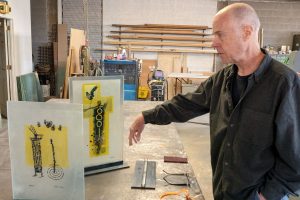 John Suttman stands beside recent glass sculptures in his workshop in downtown Winslow, where he has been experimenting with painting and firing layered glass while continuing the metalwork that defines much of his career.