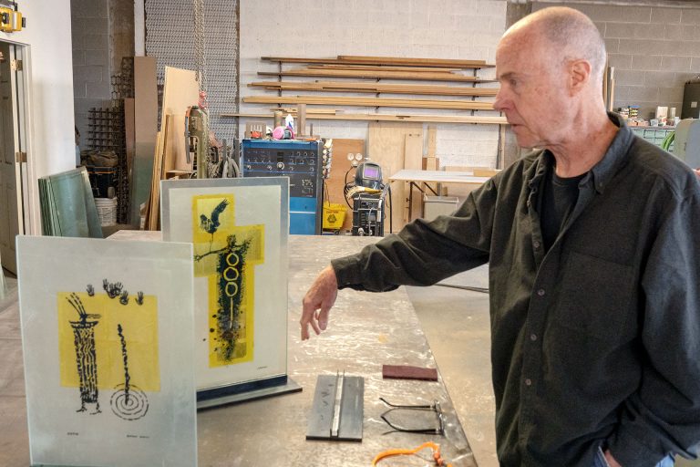 John Suttman stands beside recent glass sculptures in his workshop in downtown Winslow, where he has been experimenting with painting and firing layered glass while continuing the metalwork that defines much of his career.
