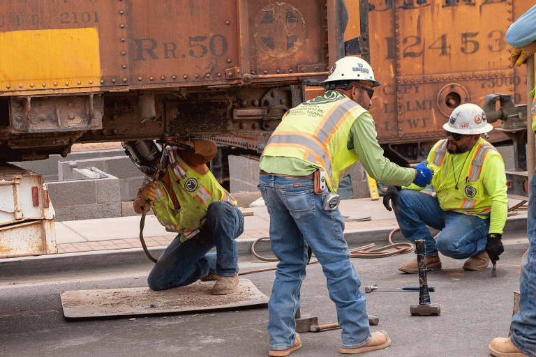Tegan Chacon (left) with McCauley Construction, welds components in preparation for installing one of the Santa Fe boxcars that will be converted into public restrooms in downtown Winslow, as Marco Villa Gomez (center) and Niver Garcia (right) stand nearby assisting with the work.