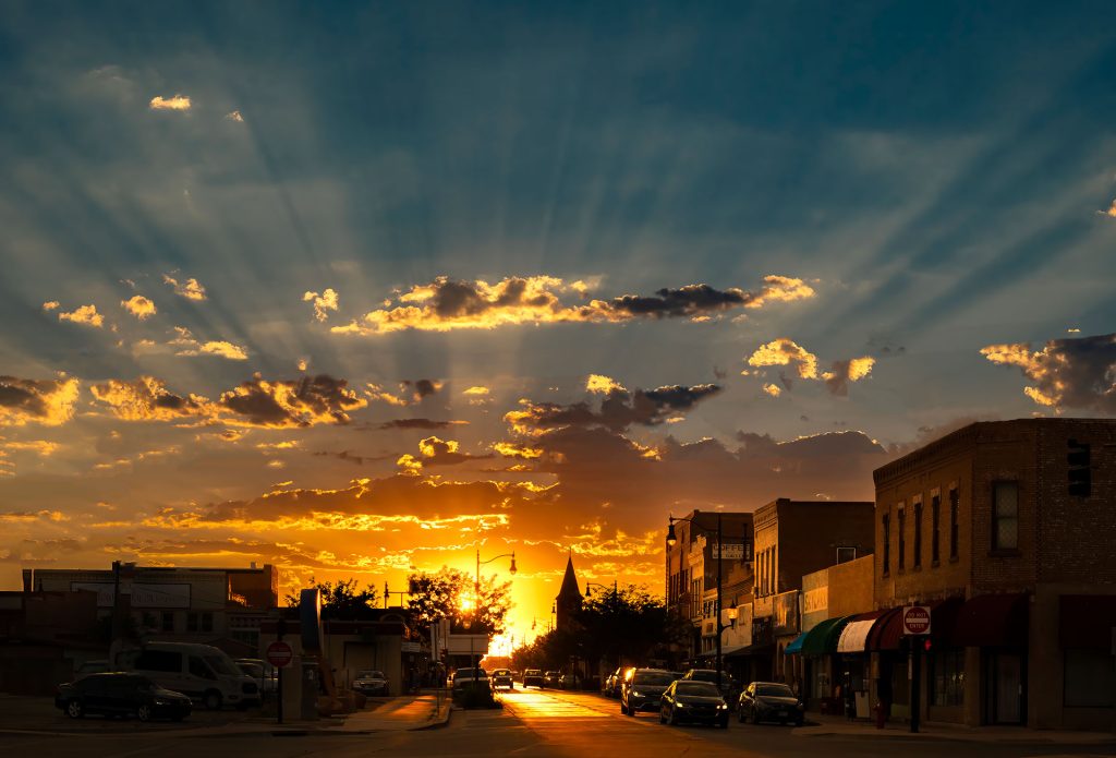 Golden light spills across downtown Winslow as the late-summer sun sinks toward the horizon, sending long shafts of light streaming over the historic storefronts and casting the street in a warm evening glow.