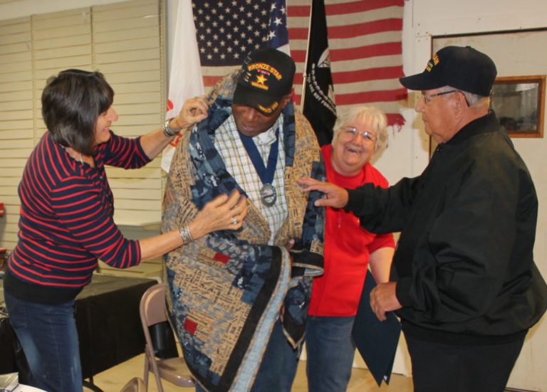 Photo by Chanae Nielson - Veteran Walter Blackman (center) is wrapped in the Quilt of Valor honoring his military service awarded to him by members of the group Heart to Heart Quilts (left to right) Bonnie Tarlton, Cheryl Voirin and Bob Bernal.