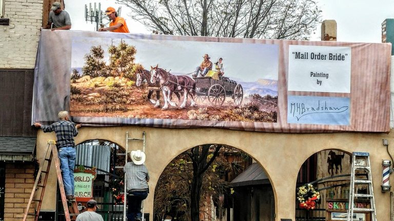 Photo by Tess KennaThis reproduction of Mel Bradshaw’s “Mail Order Bride” now hangs above the entry of the Skylark Courtyard in historic downtown Winslow. Shown here hanging the mural on the ladders are Steve Adams (left) and Lawrence Kenna (right), along with Adam’s construction crew.