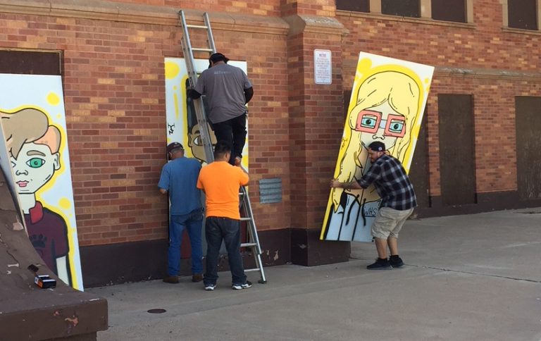 Photo by Sam CrispWinslow artist Julian Chavez (right) carries a panel of his artwork to be displayed on the exterior walls of the decommissioned high school located at Cherry Street and Apache Avenue in Winslow. Chavez has now completed eight of the 12 panels he has planned.