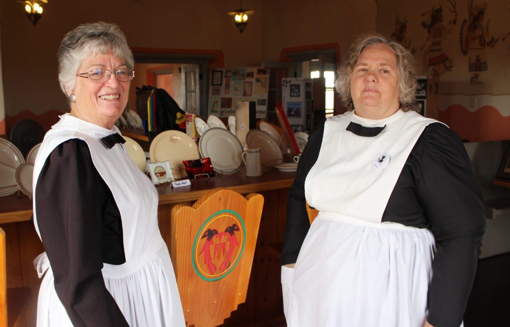 Photo by Toni GibbonsPeggy Nelson (left), president and head waitress of the Winslow Harvey Girls, stands with Linda Hallman (right) during the cultural demonstration at the Painted Desert Inn on Aug. 25. Behind the women are some of the dishes that were used at the Harvey House restaurants. The uniform is a replica of what was worn in the late 1800s, with the exception that the dresses are made of polyester instead of wool.