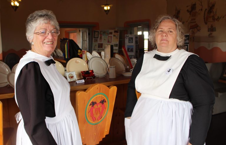 Photo by Toni GibbonsPeggy Nelson (left), president and head waitress of the Winslow Harvey Girls, stands with Linda Hallman (right) during the cultural demonstration at the Painted Desert Inn on Aug. 25. Behind the women are some of the dishes that were used at the Harvey House restaurants. The uniform is a replica of what was worn in the late 1800s, with the exception that the dresses are made of polyester instead of wool.